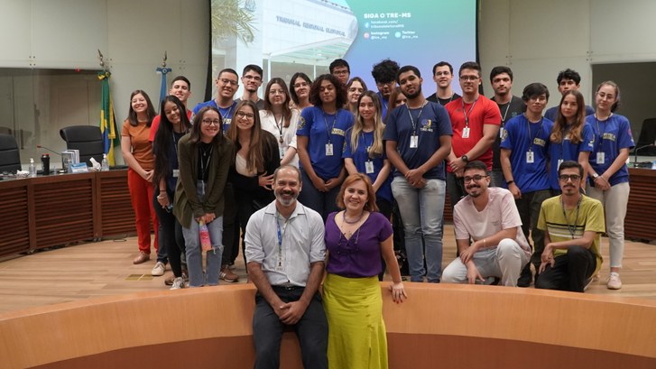 Foto dos estagiários do TRE alinhados lado a lado num palco, são homens e mulheres, vestem unifo...